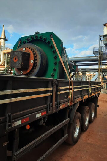 Equipamento industrial de grande porte amarrado sobre a carroceria, dentro de uma planta industrial.